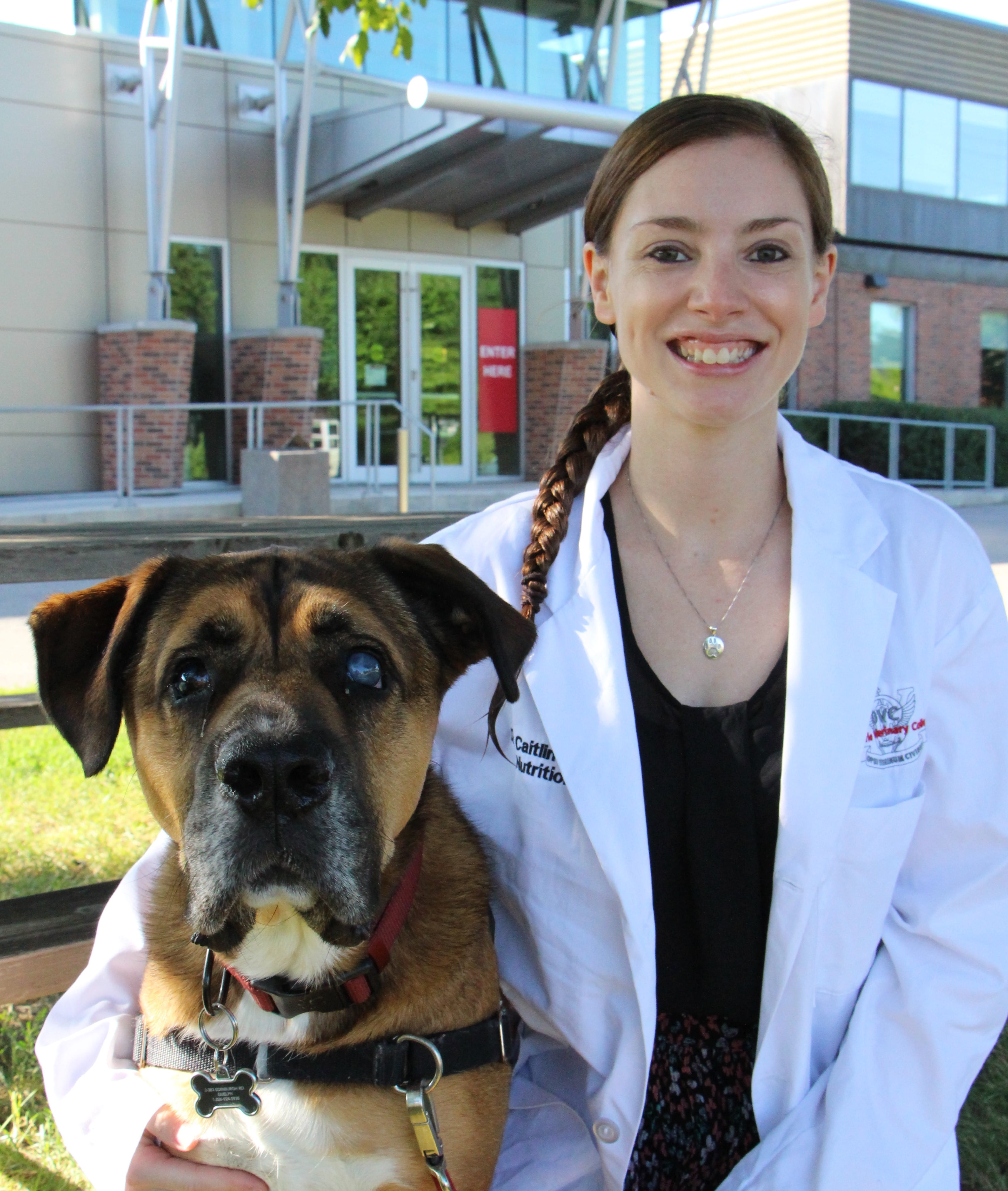 Caitlin and her dog Pilot. Caitlin is standing outside of OVC wearing a black outfit and a white lab coat. 